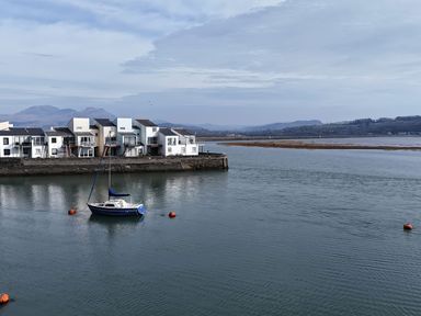 A sailboat moored in calm water near a row of white houses with mountains in the background at Fisherman Cove in Porthmadog