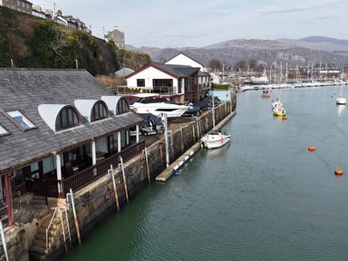 A marina with boats docked alongside buildings and hills in the background at Fisherman Cove in Porthmadog