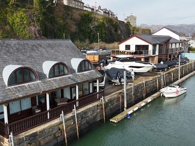 A waterfront scene with boats docked near buildings and a rocky cliff in the background at Fisherman Cove in Porthmadog