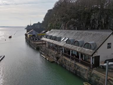 A waterfront building with arched windows along a stone quay with trees on a hill at Fisherman Cove in Porthmadog