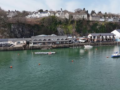 A waterfront with boats docked near buildings and houses on a hillside at Fisherman Cove in Porthmadog