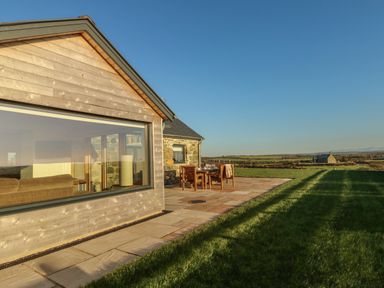 An outdoor stone patio with a wooden table and chairs next to a modern building and grass at Cuddfan in Llanfwrog near Bodedern