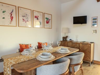 A dining room with a wooden table set with plates glasses and cutlery four gray chairs a sideboard with a lamp and a small TV on the wall at Cuddfan in Llanfwrog near Bodedern
