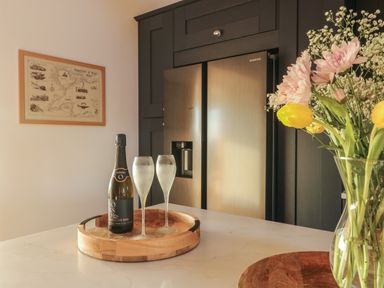 A kitchen countertop with a bottle of prosecco and two glasses on a wooden tray next to a vase of flowers and a stainless steel refrigerator in the background at Cuddfan in Llanfwrog near Bodedern