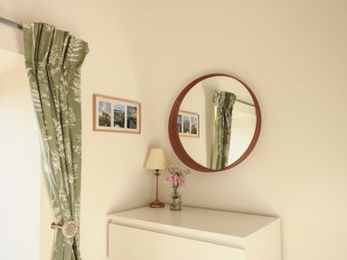 A dresser with a lamp and flowers under a round mirror with green curtains and framed pictures on the wall at Cuddfan in Llanfwrog near Bodedern