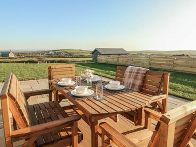 An outdoor wooden dining table set with benches and chairs set for a meal in a garden with a fence and hills in the background at Cuddfan Llanfwrog near Bodedern