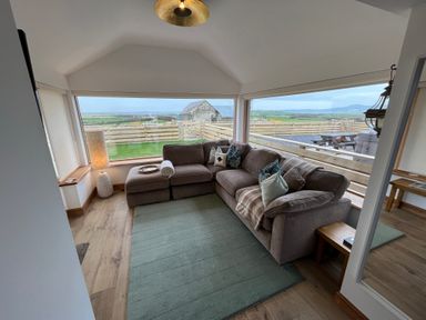 A living room with a sectional sofa and a lamp next to large windows showing a fenced yard at Cuddfan in Llanfwrog near Bodedern