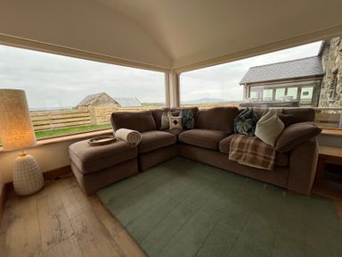 A corner seating area with a brown sofa and cushions near large windows showing a rural view at Cuddfan in Llanfwrog near Bodedern