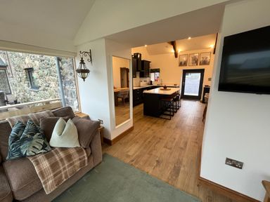 A living area with a sofa and cushions next to a window and an open kitchen with an island and bar stools at Cuddfan in Llanfwrog near Bodedern