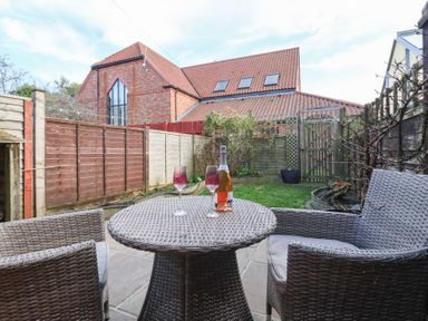 A small backyard with a round wicker table and two matching chairs on a stone patio with a bottle and two glasses on the table and houses with pitched roofs in the background