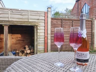 A patio table with two empty wine glasses and a bottle of sparkling rose wine on a backyard with a wood storage and wooden fences