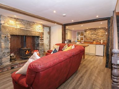 A living room with a red sofa facing a stone fireplace with a wood stove and an open kitchen with wooden countertops at Miners Rest in Blaenau Ffestiniog