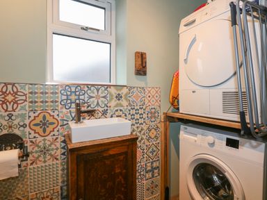 A laundry room with a sink on a wooden cabinet patterned tiles on the wall a washing machine and dryer stacked on a wooden shelf at Miners Rest in Blaenau Ffestiniog