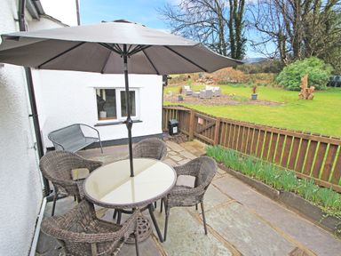 A patio with a table and four chairs under an umbrella next to a house and a garden with outdoor furniture at Igamogam in Llanllyfni near Penygroes