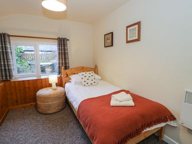 A bedroom with a single bed with an orange blanket and white towels on it a round wicker side table with a lamp plaid curtains on the window and framed pictures on the wall at Igamogam in Llanllyfni near Penygroes