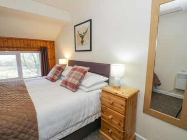 A bedroom with a double bed two plaid pillows a wooden bedside table with a lamp a window and a wall mirror at Igamogam Llanllyfni near Penygroes