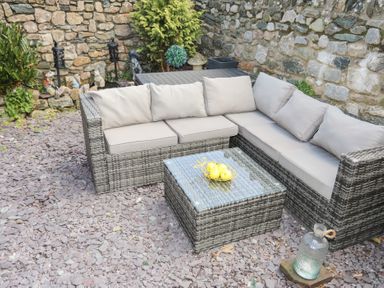 An outdoor patio area with a gray wicker sectional sofa with beige cushions a matching coffee table with a glass top a bowl with yellow apples on it stone walls and ground covered with purple slate stones at Camwy in Brynsiencyn