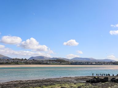 A coastal view with water and rocks in the foreground a town with buildings and hills in the background under a blue sky at Camwy in Brynsiencyn