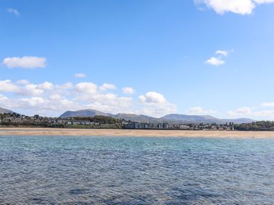 A body of water with a sandy shore and buildings with hills in the background at Camwy in Brynsiencyn