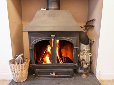 A lit wood-burning stove with a basket of newspapers on the left and fireplace tools on the right at Ty Glyn in Beddgelert