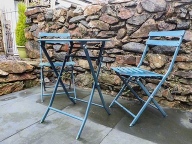 A small outdoor patio area with a blue metal table and two blue metal chairs in front of a stone wall at Ty Glyn in Beddgelert