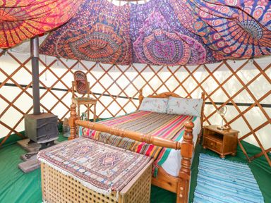 A wooden bed with striped bedding inside a yurt with a wood stove a wooden chair a nightstand with a lamp and patterned fabric on the ceiling at Brandon House Farm Yurt Brandon Parva near Mattishall