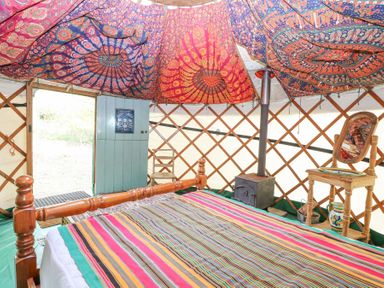 A yurt interior with a wooden bed with striped bedding a small wood stove a wooden chair with a mirror and colorful fabric hanging from the ceiling at Brandon House Farm Yurt Brandon Parva near Mattishall