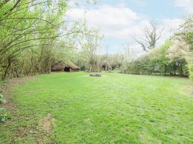 A grassy yard with small thatched huts and a fire pit surrounded by trees at Brandon House Farm Yurt in Brandon Parva near Mattishall