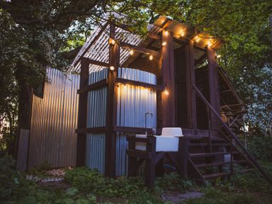 An outdoor structure with corrugated metal walls wooden frame string lights and a sink surrounded by trees and plants at Brandon House Farm Yurt Brandon Parva near Mattishall