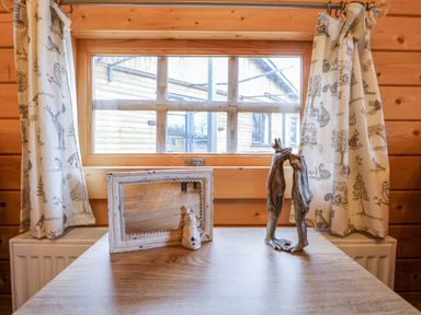 A wooden table under a window with animal patterned curtains and decorative items including a white framed mirror and a metal frog sculpture at Enchanted Cottage in Snettisham