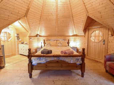 A bedroom with a wooden bed, rolled towels on the bed, wooden walls and ceiling, a cushioned chair and storage cabinets at Enchanted Cottage in Snettisham