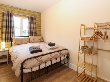 A bedroom with a bed linens with deer designs a wooden side table lamp curtains and a wooden shelving unit with hangers at Rabbit Warren Lodge in Snettisham