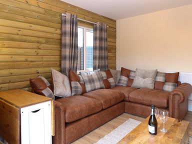 A corner sofa with cushions next to a window with curtains and a wooden table with a bottle and glasses at Robins Nest Lodge in Snettisham