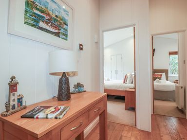 A hallway with a wooden table holding a lamp and decorations leading to two bedrooms each with a bed and pillows at 1 Miller's Island near Lanreath
