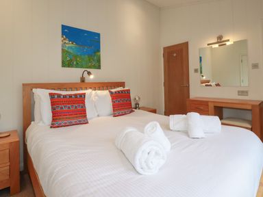 A bedroom with a double bed with rolled white towels and colorful pillows a wooden door and a dresser with a mirror at 1 Miller's Island near Lanreath
