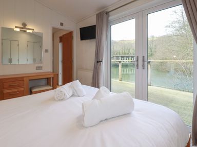 A bedroom with a bed and rolled towels on it a wooden desk with mirror a mounted television and a glass door overlooking a water body at 1 Millers Island near Lanreath