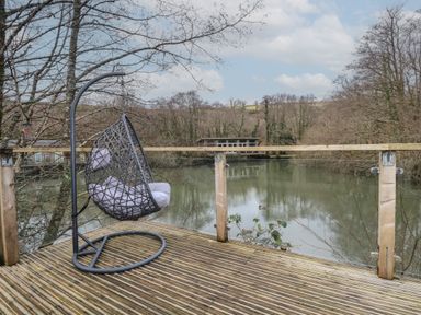 A wooden deck with a hanging chair overlooking a river with trees and a house in the background at 1 Miller's Island near Lanreath