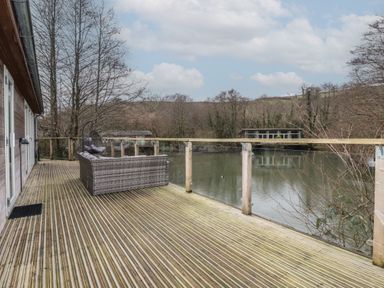 A wooden deck with outdoor seating overlooking a lake and trees at 1 Millers Island near Lanreath