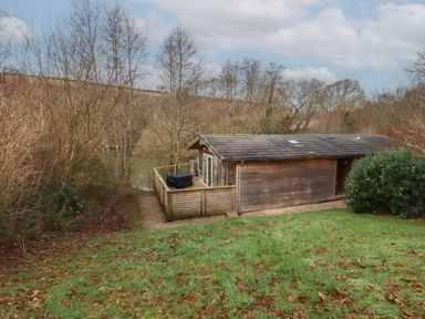 A wooden cabin with a deck and outdoor grill surrounded by trees and grass at 1 Miller's Island near Lanreath