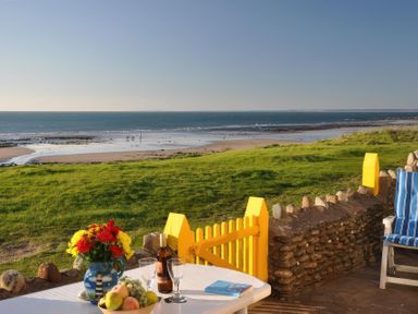 A patio with a table holding flowers fruit and wine glasses overlooking a grassy area and a beach at Homestead Aberdesach near Penygroes