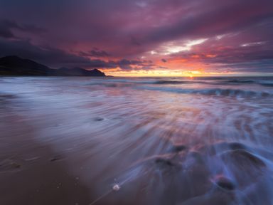 A beach with waves and a sunset sky with clouds and mountains in the background