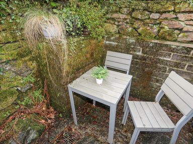 A small outdoor corner with a gray table and two chairs a potted plant on the table and plants growing on mossy stone walls at Quarrymans cottage in Beddgelert