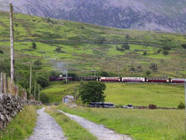 A countryside landscape with a gravel path a train on railway tracks and mountains in the background at Quarrymans cottage in Beddgelert