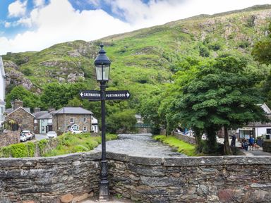 A stone bridge over a river with a lamppost and directional signs in a village with hills in the background at Quarryman's cottage in Beddgelert