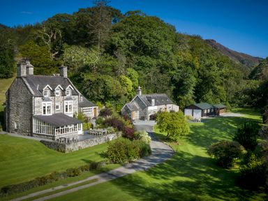 A stone house with multiple chimneys and an adjoining building surrounded by trees and lawn at Tan Llan in Llanelltyd near Dolgellau