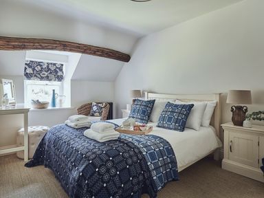 A bedroom with a bed with blue patterned bedding and pillows two bedside tables with lamps a window with floral blinds and a wicker chair at Tan Llan in Llanelltyd near Dolgellau
