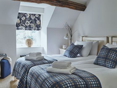 A bedroom with two single beds with blue patterned blankets and pillows a wooden beam and a window with floral blinds at Tan Llan in Llanelltyd near Dolgellau
