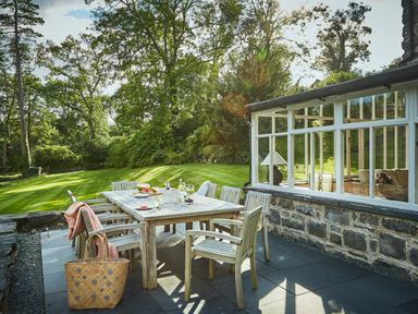 An outdoor dining area with a wooden table and chairs on a stone patio next to a glass conservatory at Tan Llan in Llanelltyd near Dolgellau