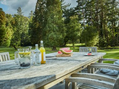An outdoor wooden table with bottles and sliced watermelon on a cutting board surrounded by wooden chairs at Tan Llan in Llanelltyd near Dolgellau
