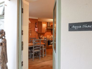 View through an open door into a kitchen dining area with wooden chairs and table and kitchen cabinets at The Aqua Shack in Newquay